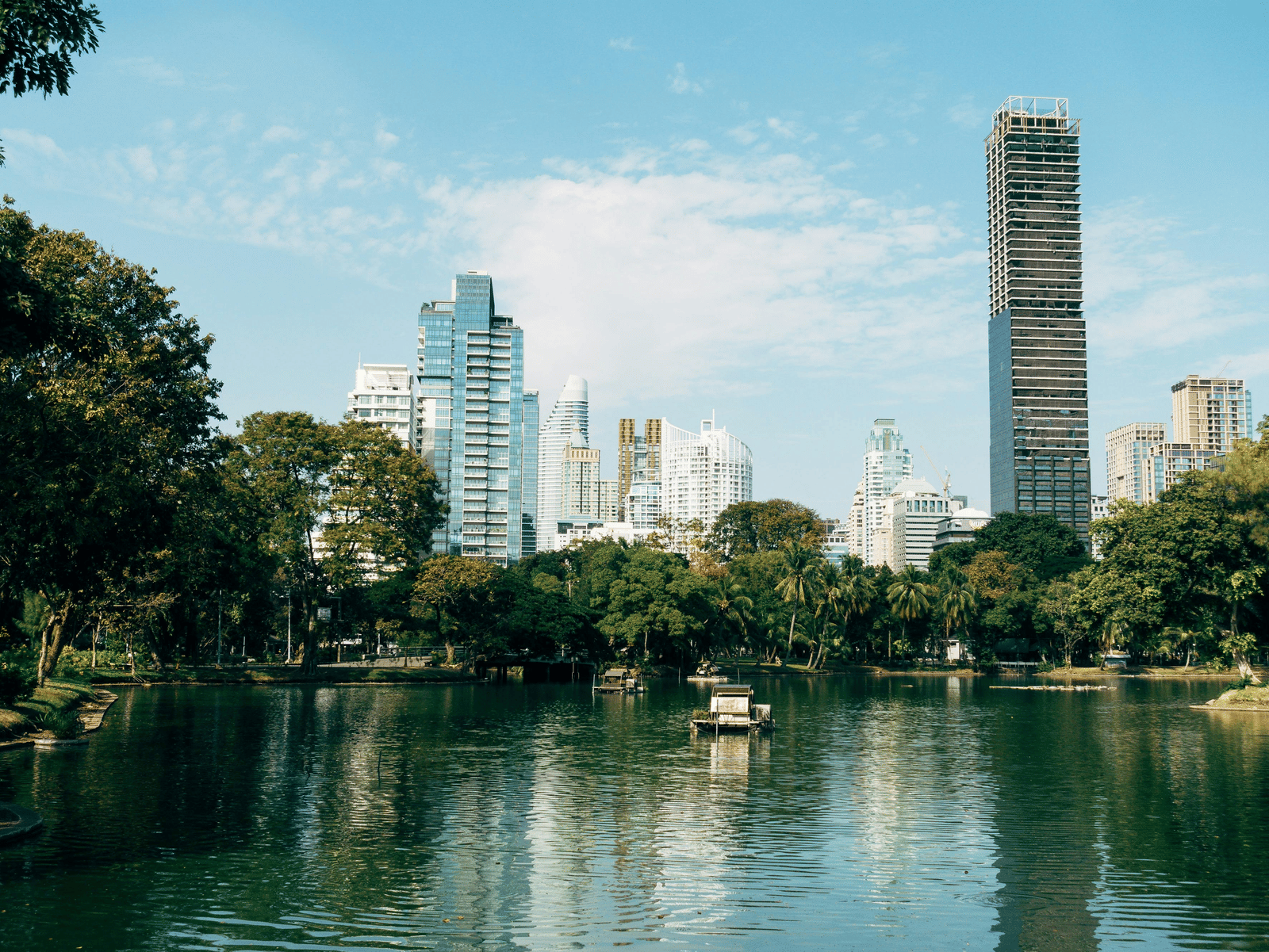 A scenic view of a city park with a calm lake in the foreground, lush green trees lining the water, and modern high-rise buildings, including a prominent thin white tower, in the background under a blue sky.