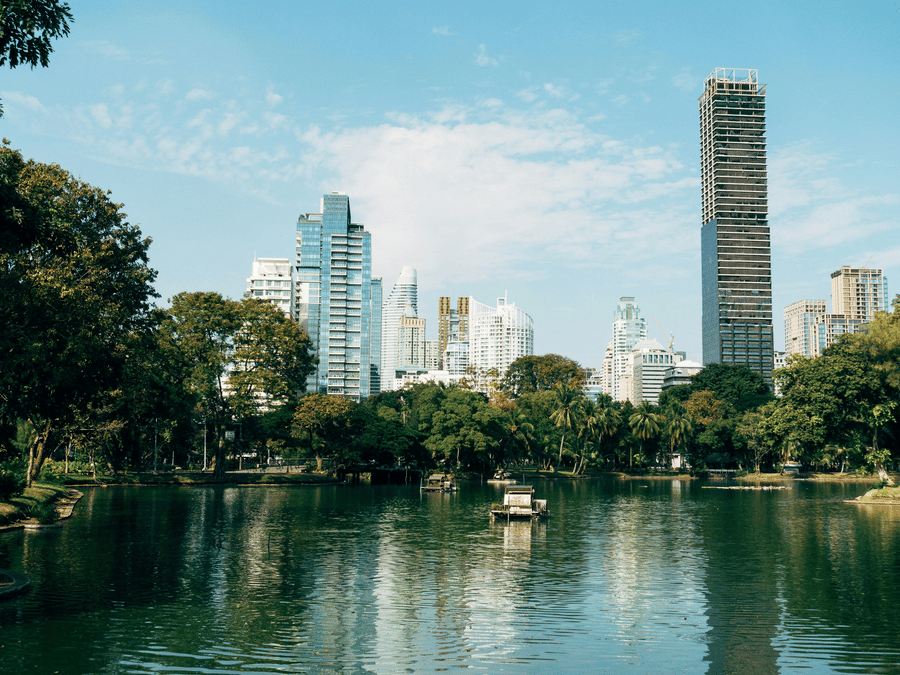A scenic view of a city park with a calm lake in the foreground, lush green trees lining the water, and modern high-rise buildings, including a prominent thin white tower, in the background under a blue sky.