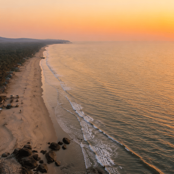 Aerial view of Mandrem Beach at sunset with palm-lined sand, thatched beach huts, waves rolling in, and the sun dipping toward the Arabian Sea.