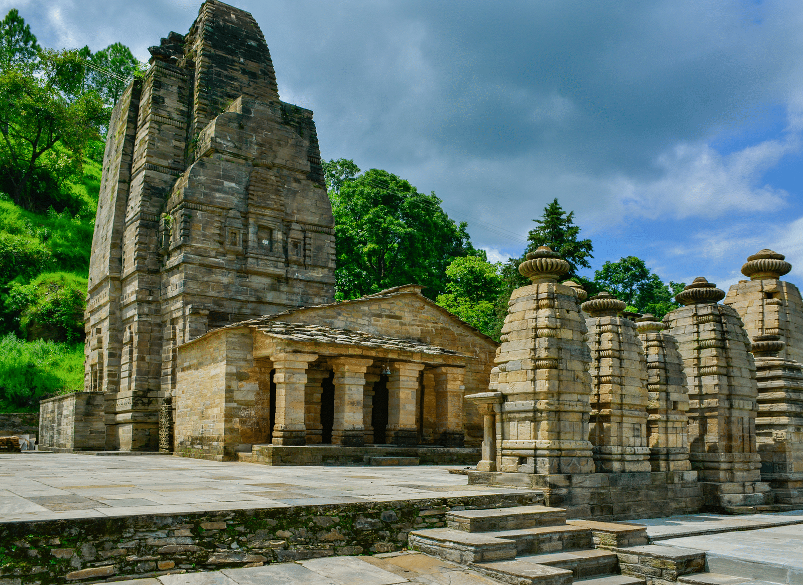 Ancient stone temple complex surrounded by greenery under a cloudy blue sky, identified as Katarmal Temple.