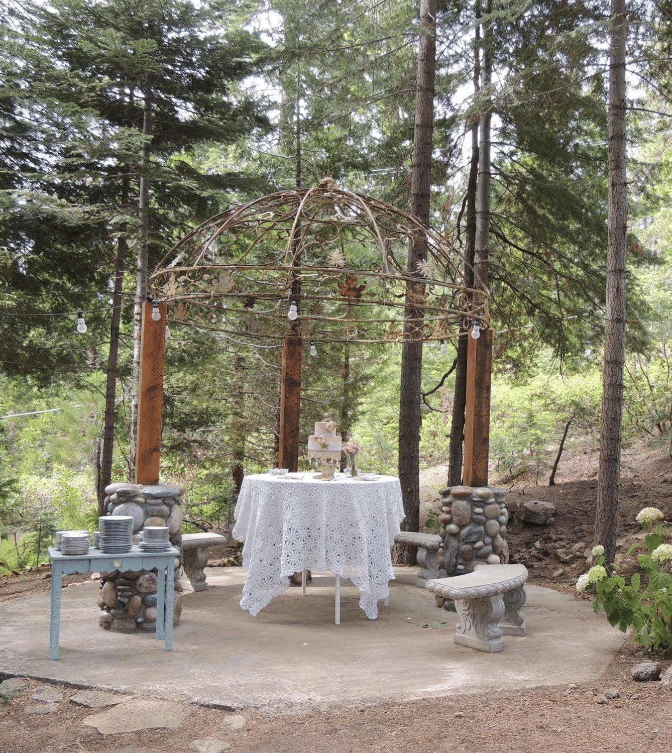 A wedding cake placed on a table in the middle of a forest with a stack of plates placed on a table beside it