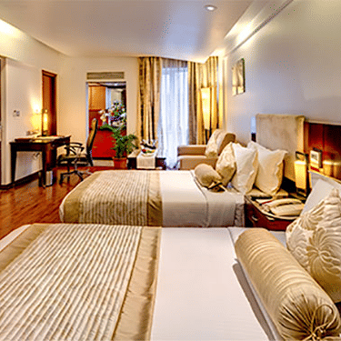 Side view of a Premium Room at Hotel Hindusthan International Select Bengaluru with twin beds, wall-mounted TV, wooden flooring, and warm ambient lighting.