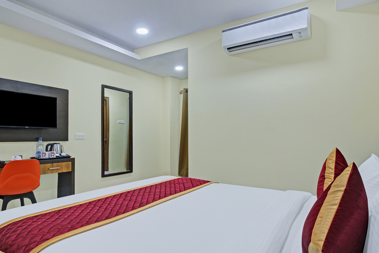 A hotel bedroom with double bed featuring white linens and red accent pillows, a black upholstered headboard, a mirror, an orange study chair and a simple, uncluttered decor.