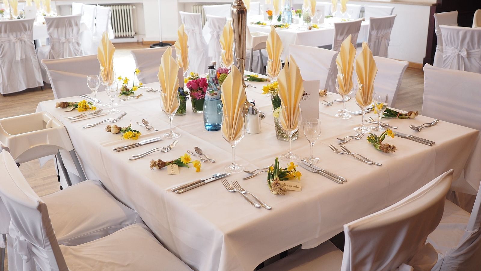 A bright and airy dining area with tables set with white linens, golden yellow napkins and colourful spring flowers in small vases, creating a cheerful ambience
