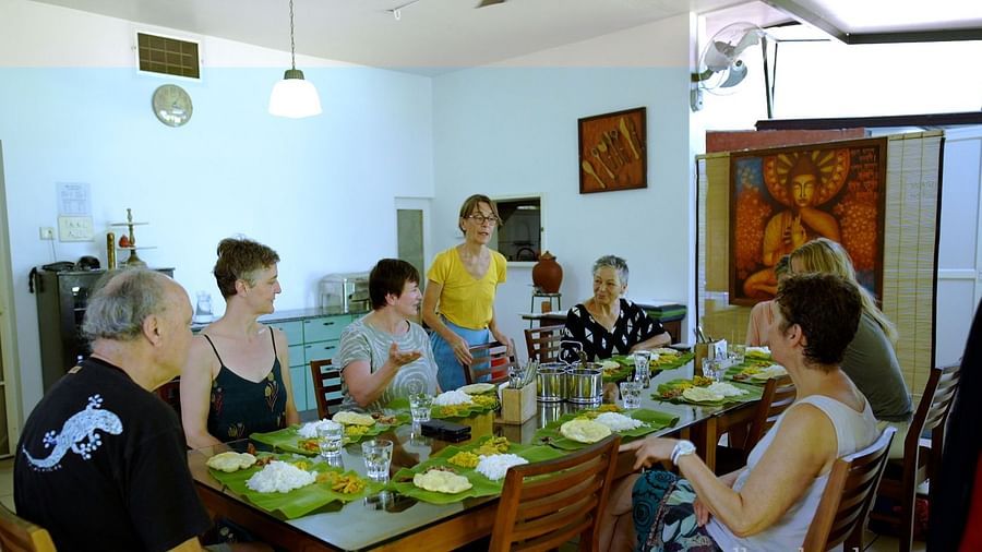 A group of people are seated around a long table indoors, sharing a meal served on banana leaves, with one person standing.