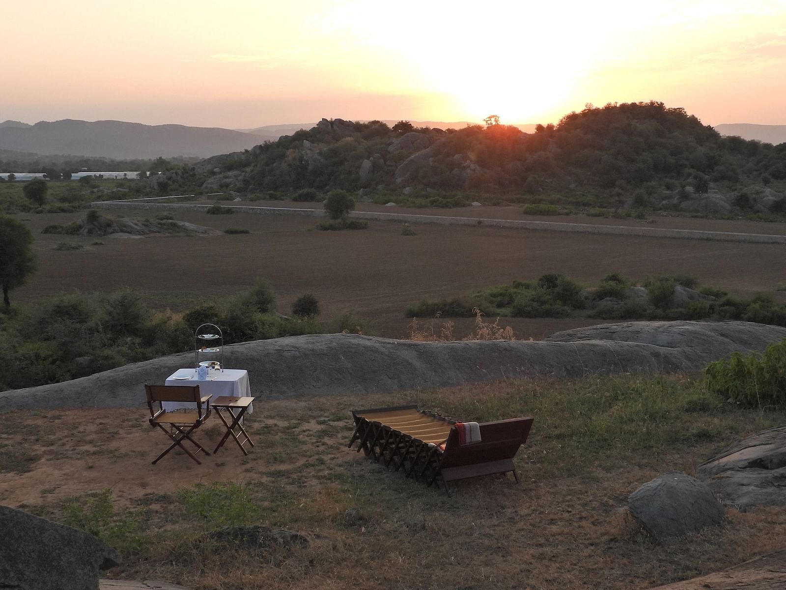 An image of camping site where a chair and table is placed near a lounger bed, and the sun is also setting at the horizon - Utsav Camp Sariska