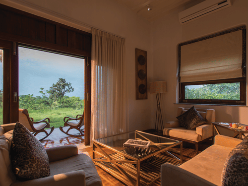 Sun illuminating the interiors of a room at The Serai Bandipur, one of the best Romantic Getaways near Bangalore, featuring a sofa set and a patio seen through the doorway with a couple of chairs facing the trees outside