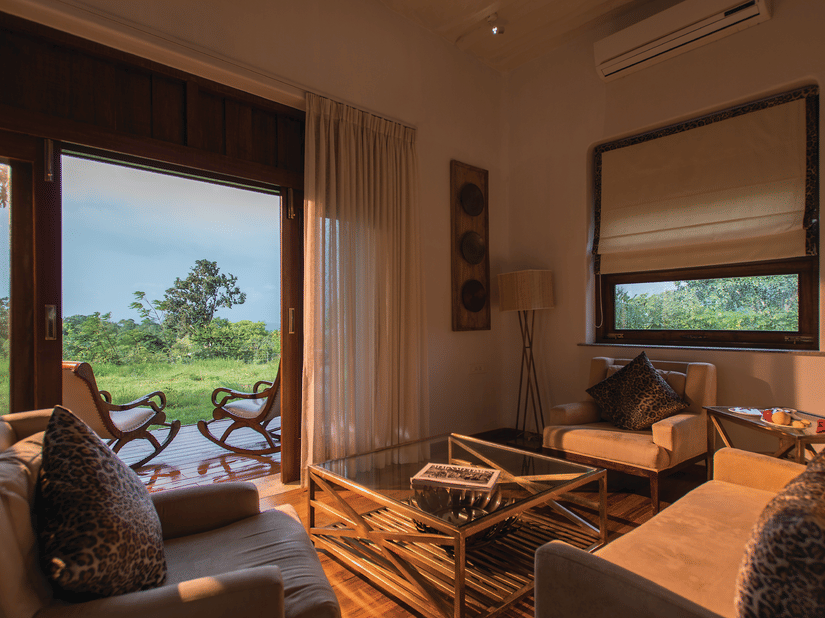 Sun illuminating the interiors of a room at The Serai Bandipur, one of the best Romantic Getaways near Bangalore, featuring a sofa set and a patio seen through the doorway with a couple of chairs facing the trees outside