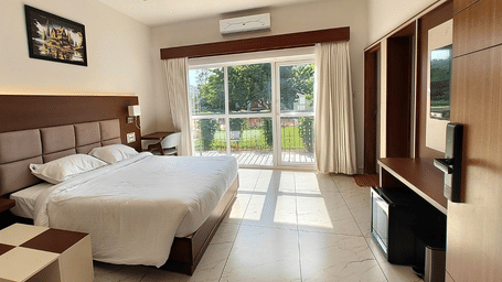 A hotel bedroom featuring a double bed, wooden furniture, a wall-mounted picture frame, and large sliding doors looking out towards greenery | Lords Sahyadri Resort, Shivamogga