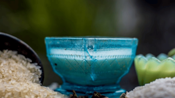 Rice and rice flour are scattered next to a glass bowl containing a white face mask or paste