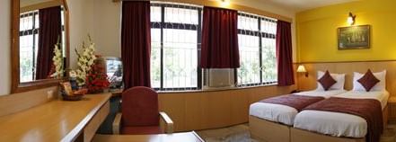 A bedroom with twin beds, a bedside table with a lamp, a chair, a desk, a console table, a mirror and large windows at Hotel Mamallaa Heritage, Mahabalipuram.