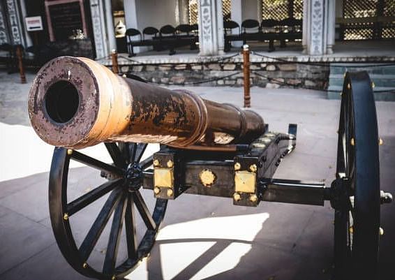 A vintage cannon rests on a wheeled carriage, positioned in front of a building with seating.