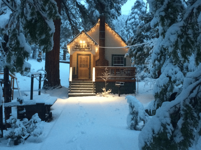 Snow covered cabin at Shaver Lake Village Hotel, with a lit doorway and porch, nestled among snow-laden pine trees.