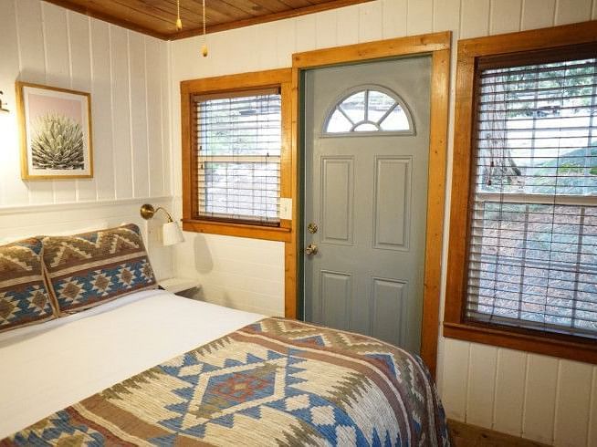 Overview of a Room at Shaver Lake Village Hotel with a bed featuring a patterned blanket, a grey door with a half-circle window, and two windows with blinds.