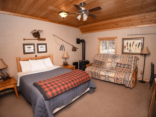 a Room with a bed, plaid blanket, patterned couch, wood ceiling, fishing decor, and two framed pictures at Shaver Lake Village Hotel.