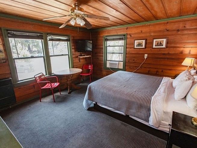 a Cozy bedroom at Shaver Lake Village Hotel with wood paneled walls, two windows, a double bed, and red chairs next to a small table.