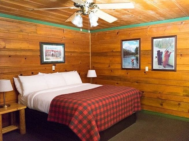 a Bedroom at Shaver Lake Village Hotel with wooden walls, plaid comforter, white pillows, framed artwork, and a ceiling fan.