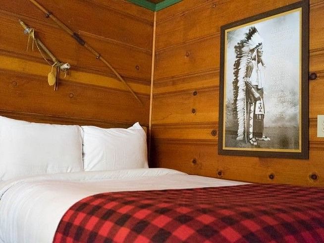 Overview of a Bed in a room at Shaver Lake Village Hotel with white pillows, red plaid blanket, wood paneled walls, framed art, and decorative sticks.