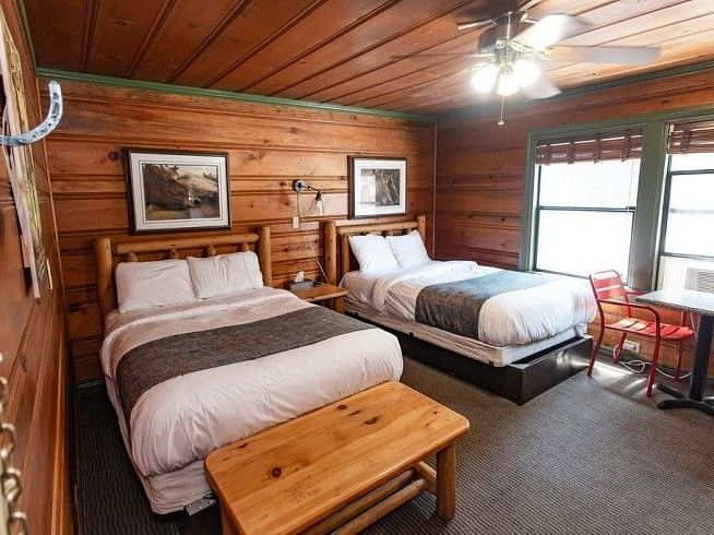Overview of a Room at Shaver Lake Village Hotel with two twin beds, wood panelled walls, a wooden table, and red chairs by a window.