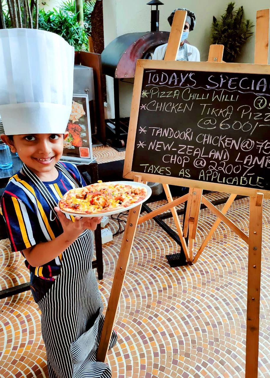 a child showing a pizza after making it in front of a signboard - Caravela Beach Resort Goa