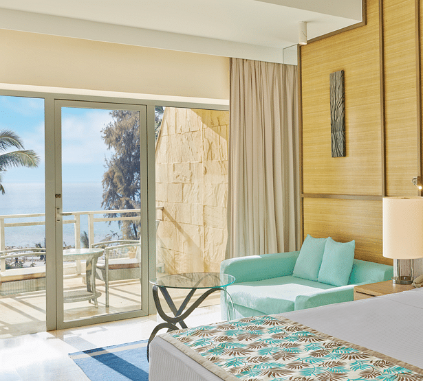 Bright, airy room with a mint-green sofa, glass coffee table, and a balcony offering a clear view of the sea and palm trees – The Resort, Mumbai