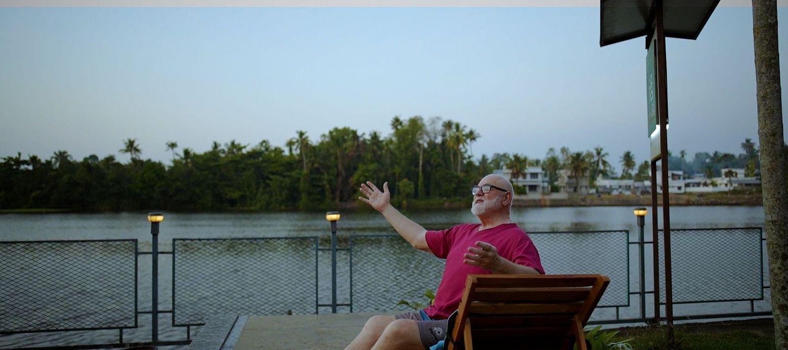 A man with white hair and a beard sits in a lounge chair by a lake, giving a thumbs-up gesture.