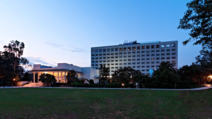 A large, multi-storey building of Clarks Amer, Jaipur stands illuminated against a deep blue twilight sky, fronted by a green lawn.