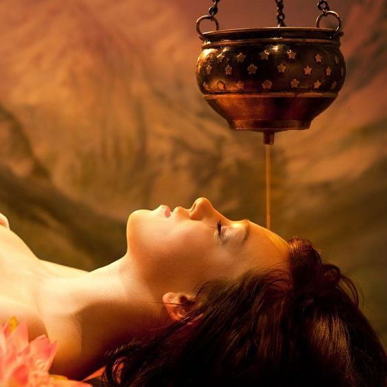 A serene woman is receiving an Ayurvedic Shirodhara treatment, with warm oil pouring from a brass pot onto her forehead.