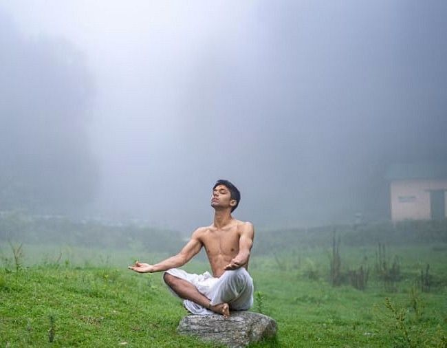 Man meditating outdoors on a grassy field surrounded by mist and trees, embracing serenity and connection with nature in a peaceful setting.