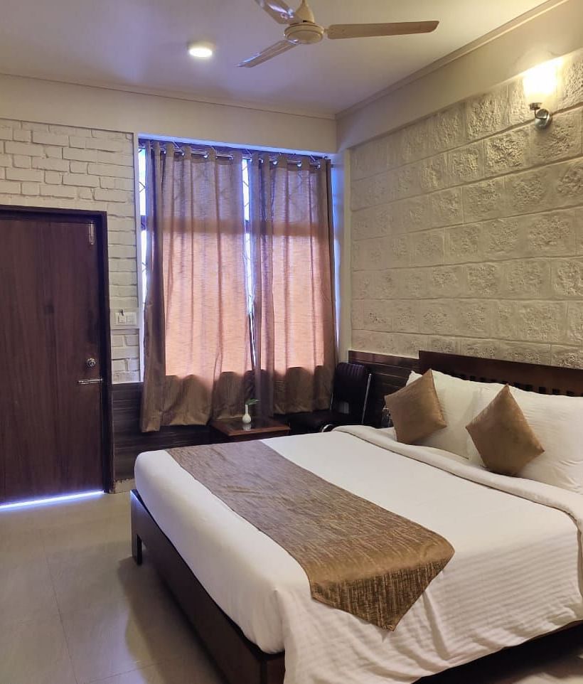 Bedroom with a huge bed, bedside table, windows with curtains, wooden door, and a TV | Standard Room | Woodstock Resorts, Coorg