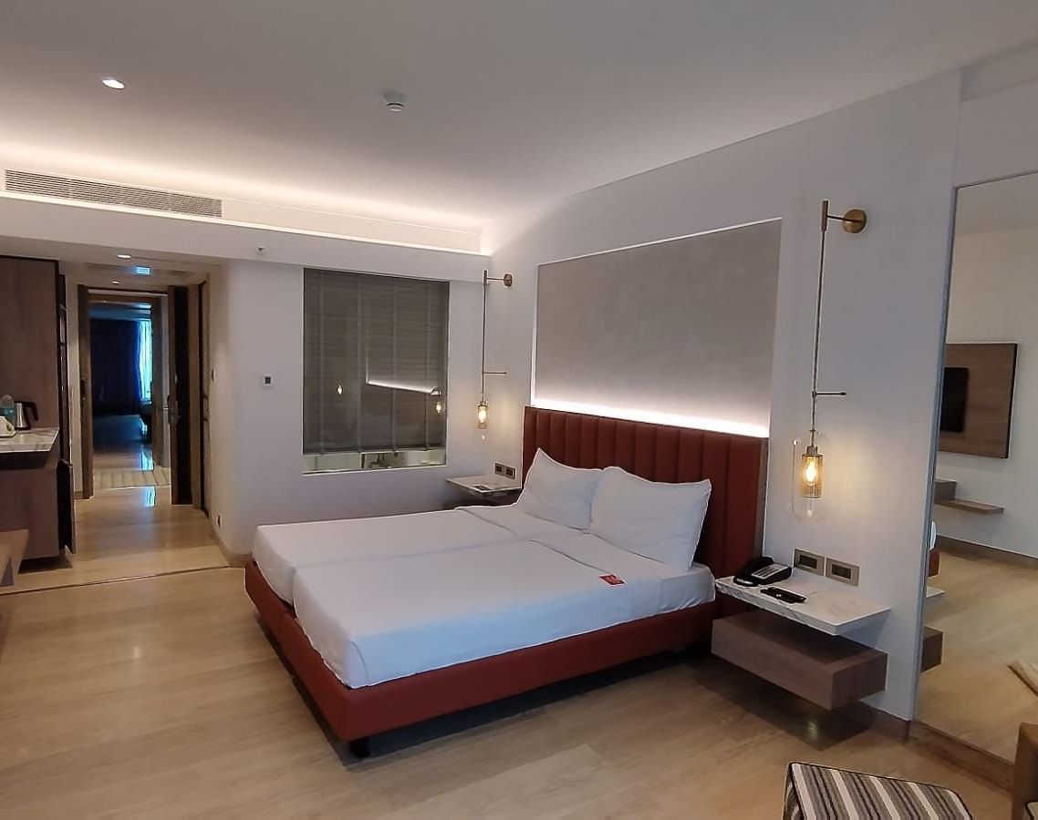 A warmly lit executive bedroom in Mirage Hotel, featuring twin beds, a large mirror, lounge chairs, and an entertainment unit.