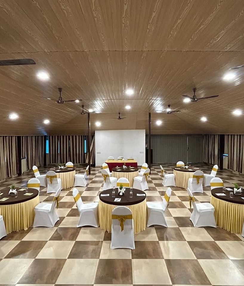 An indoor event space is arranged with round tables set with ceiling fans and lighting visible | Ballroom | Woodstock Resorts, Coorg