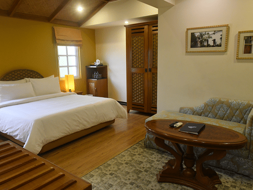 Superior Room at The Richmond, Puducherry featuring a plush bed along with a lamp next to it and a sitting area at the other end of the room with a tiny sofa and a wooden coffee table placed on a carpet