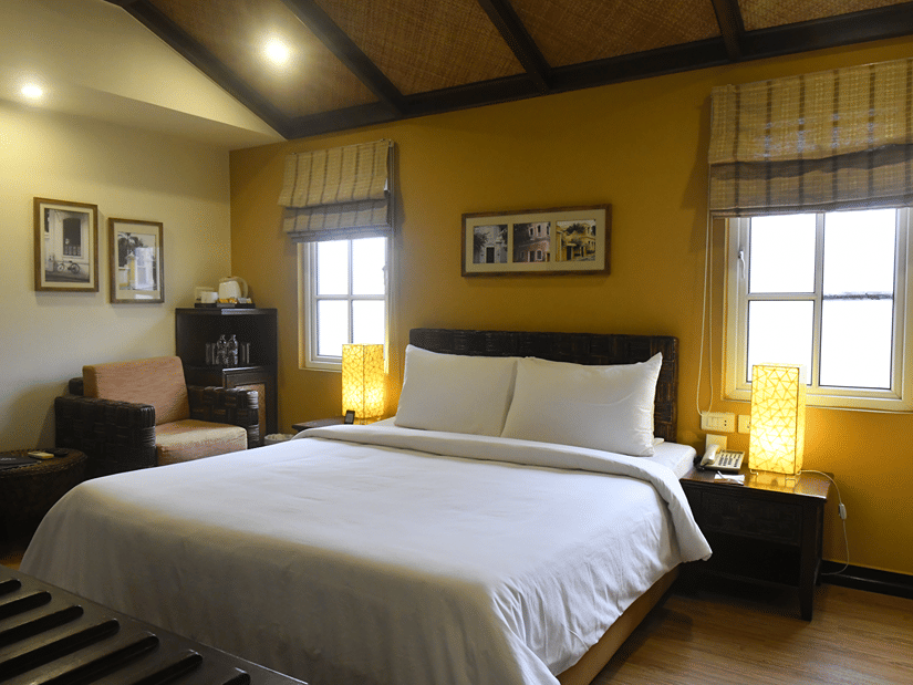 Deluxe Room at The Richmond, Puducherry featuring a plush bed with fresh linen and two lamps placed on the both sides on the nightstand as the sunlight makes its way through the two windows next to a sitting space