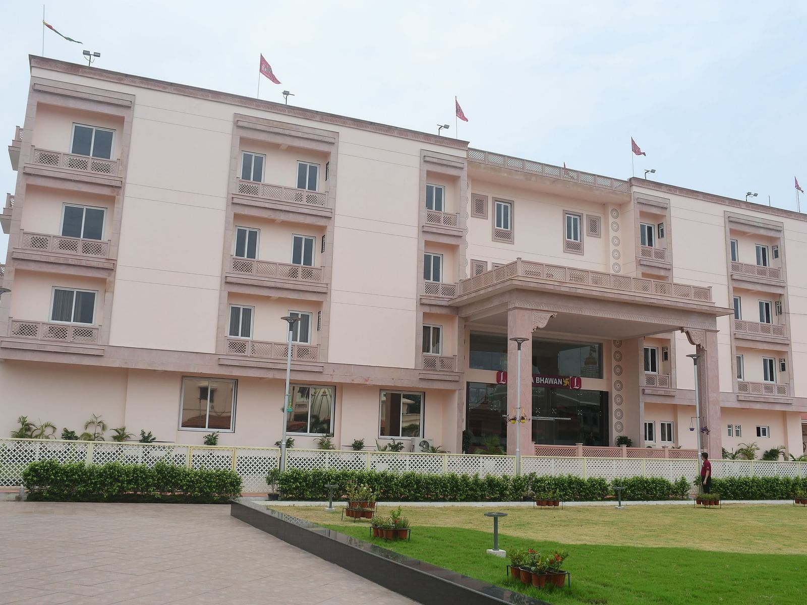 The front view of a modern, light-coloured hotel building with a covered entrance and green lawns in front is presented - Kankidham Lords Inn