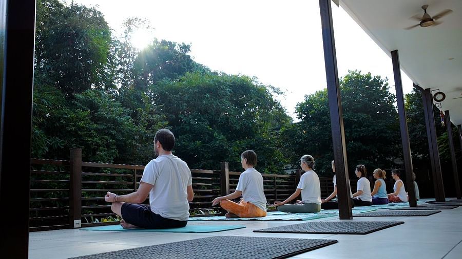 A person with short hair sits in a yoga pose outdoors, facing away from the camera, with trees in the background.