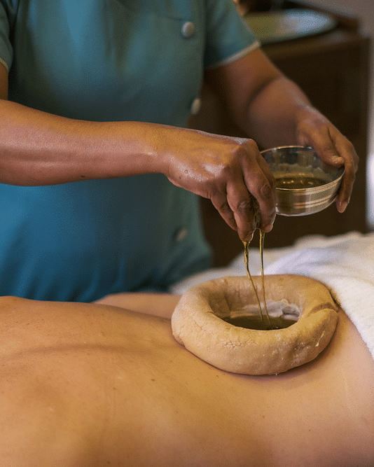 a massage therapist pouring oil into a vessel kept on the back of a client