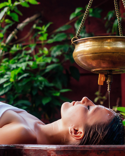 a woman lying on a spa bed with her eyes closed undergoing shirodhara therapy