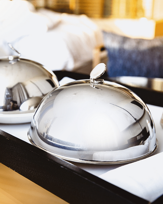 A black tray on a bed holds two covered silver dishes, a spoon on a white napkin, and salt and pepper shakers.