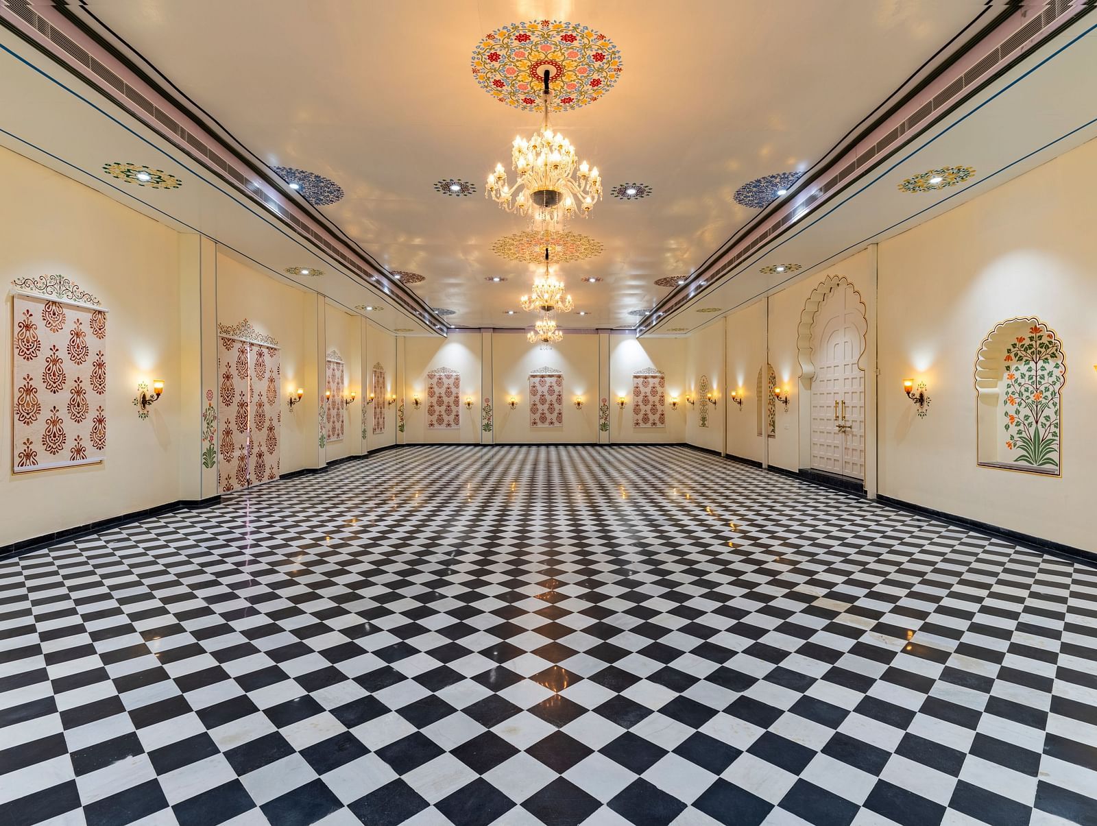 Long hallway with black-and-white checkered flooring, ornate golden chandeliers, and framed artwork on cream walls.