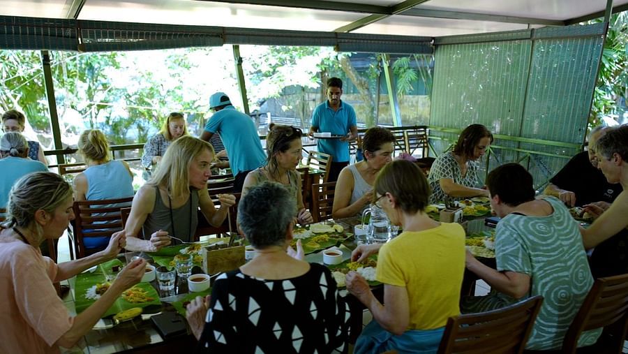 A large group of people are seated at a long table outdoors, sharing a meal and interacting with servers.