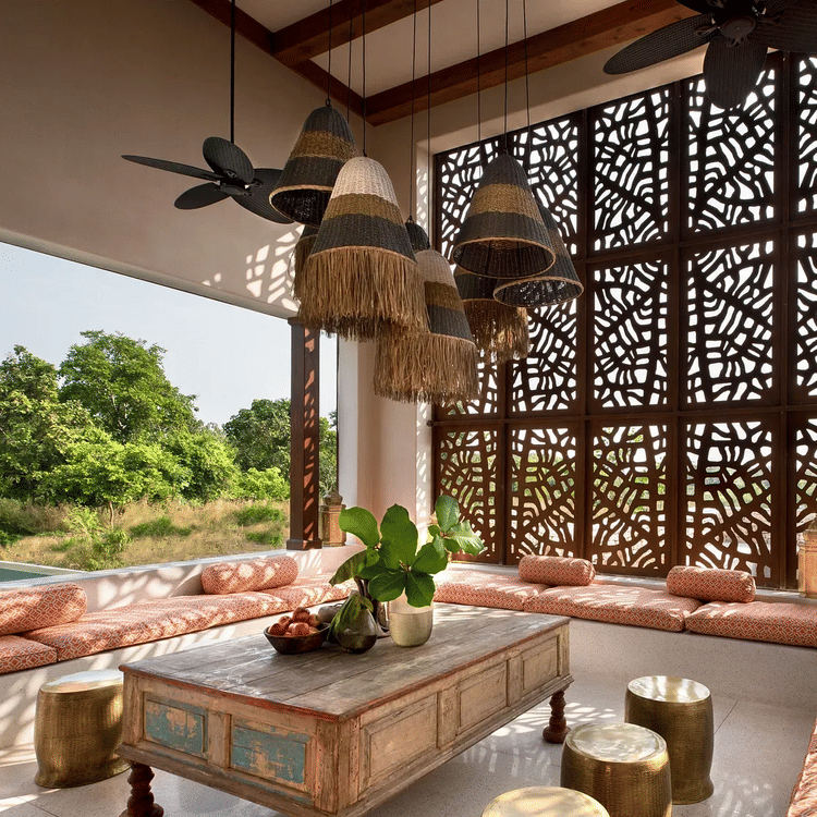 A covered outdoor lounge area with cushioned seating arranged around a low wooden table. Decorative pendant lights hang from the ceiling, and a patterned screen provides partial enclosure. Lush greenery and a body of water are visible in the background.