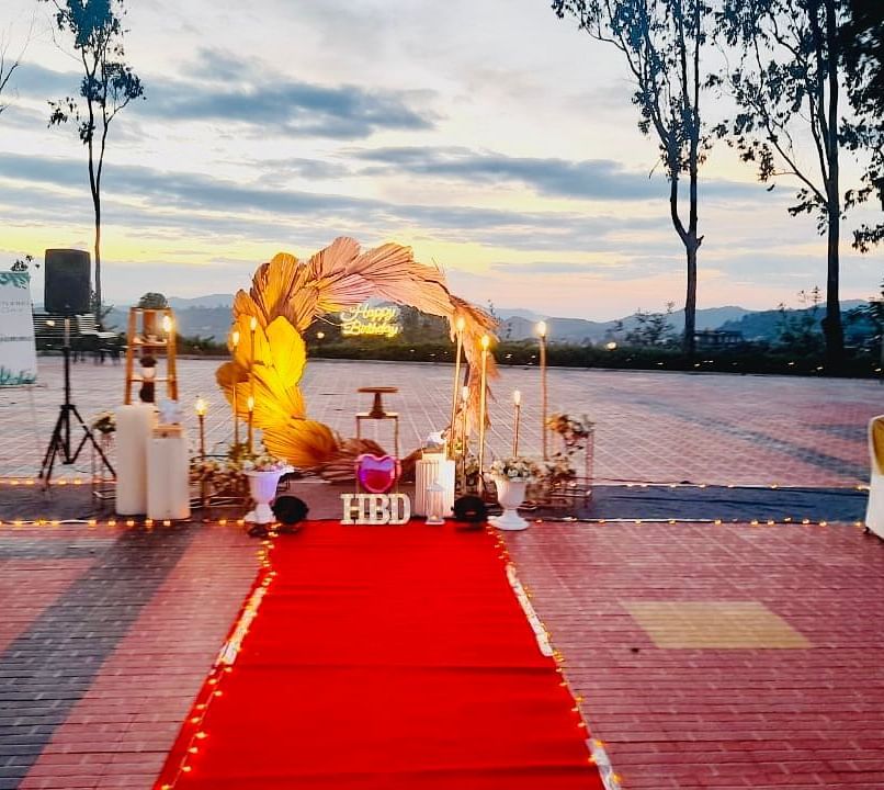An outdoor wedding setup features a red carpet leading to a floral archway, with chairs arranged on either side and greenery in the background | Open Deck | Woodstock Resorts, Coorg