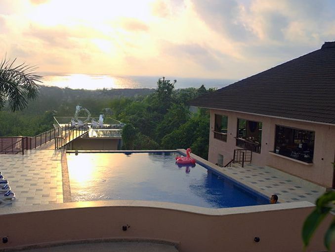 nfinity pool at Crescent Resort, Goa with sunset sea view, a pink flamingo float, lush greenery, and poolside seating under soft evening sky.