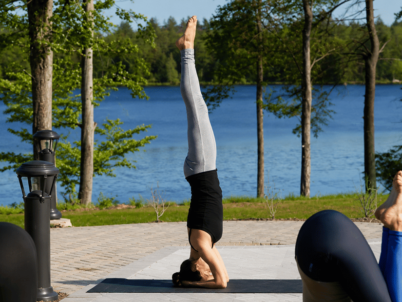 A person in a yoga headstand on a mat beside a lake, with others stretching in the foreground
