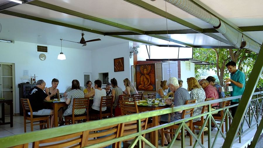 A group of people are seated at a long table indoors, sharing a meal in a bright, open space with an outdoor patio.