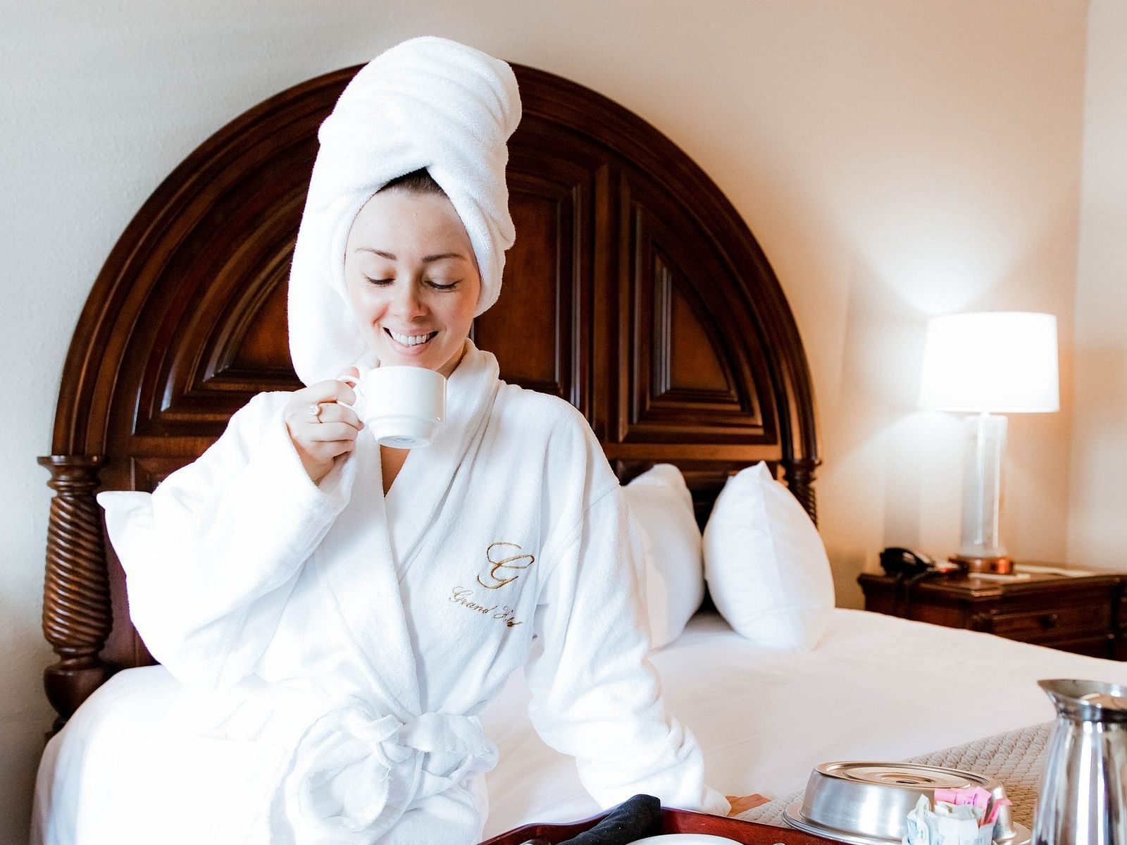 Woman in bathrobes and a towel wrapped around her head enjoying a beverage through a cup sitting on the bed with a lamp at the background and beverage containers at the corner