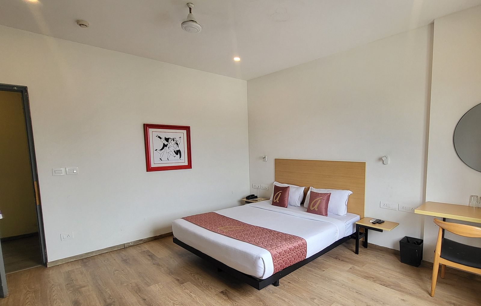 A spacious hotel room with a minimalist design, wooden flooring, a double bed, a desk, and a large circular mirror - Astra Hotels & Suites, Sarjapura