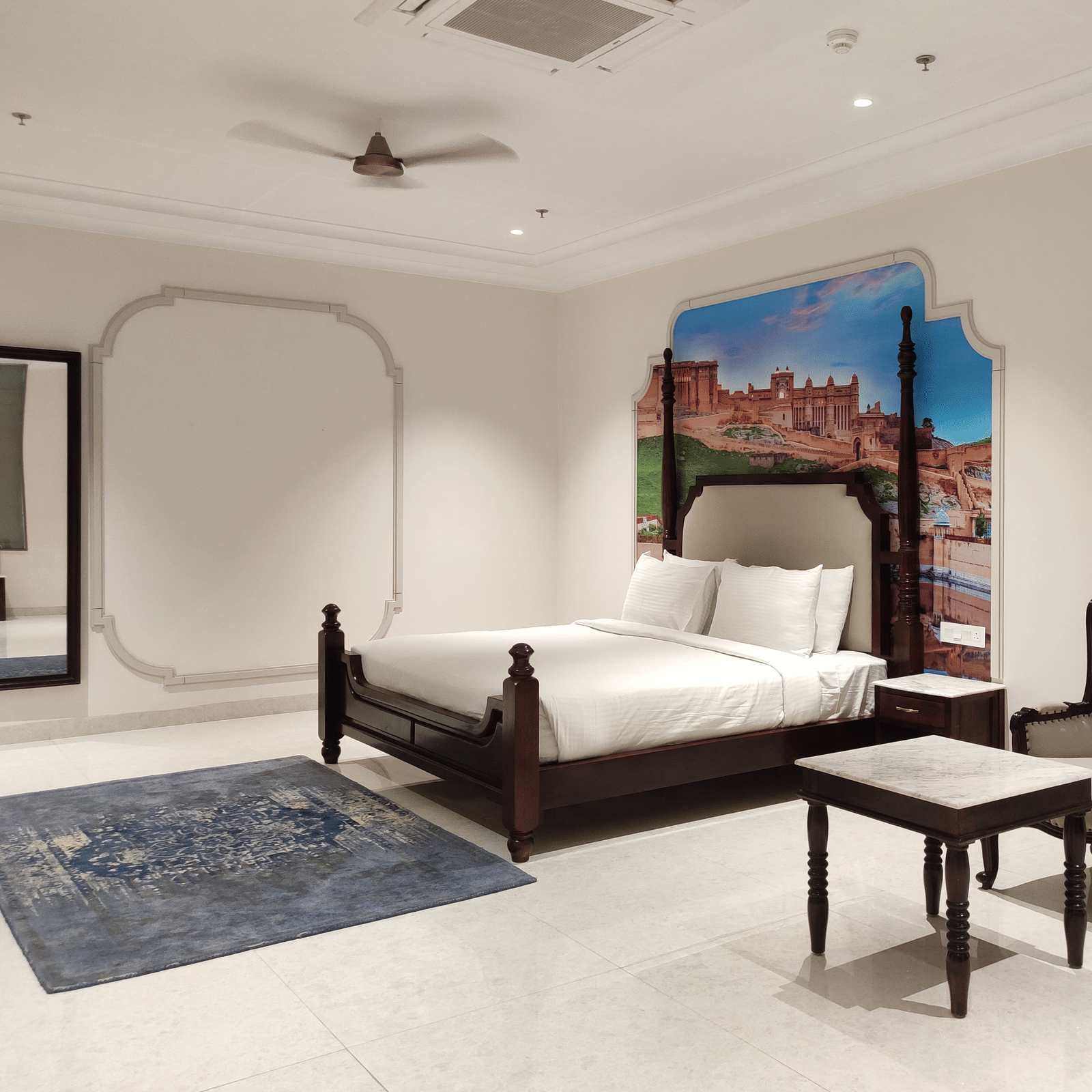 A bedroom view at Anant Mahal, Jaipur featuring a king size bed, a blue rug, a mirror, a seating area with a small table and a cushioned chair.