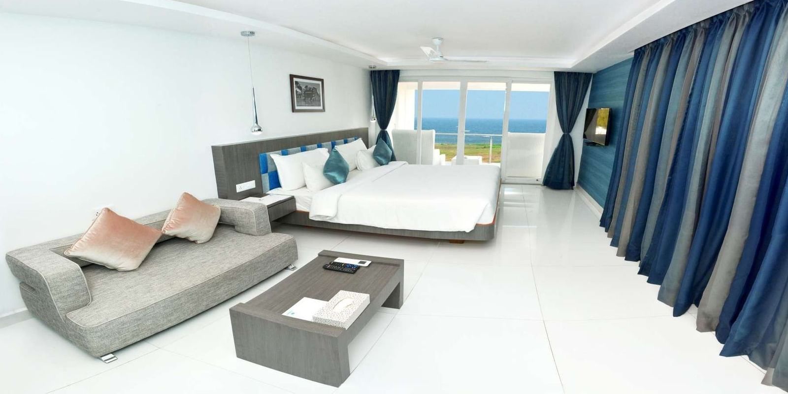 A modern hotel room with a large bed, a grey sofa, and a coffee table, featuring floor-to-ceiling windows with blue curtains offering a view of the ocean | Grande Chalet Sea View | Grand Bay Resort and Spa